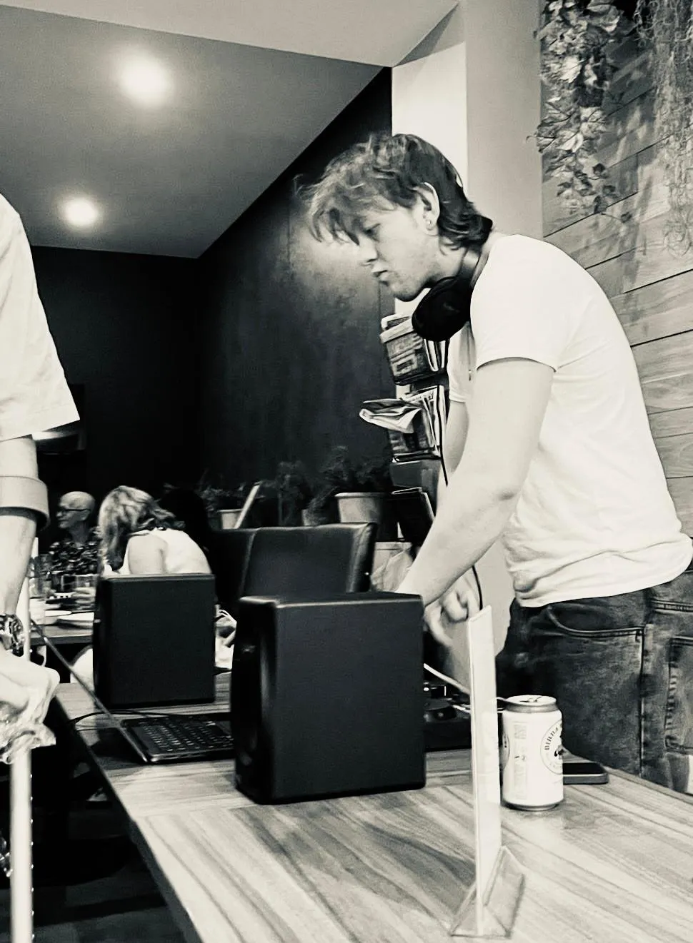 C@M in side profile at a café gig, headphones round his neck, mixing behind a pair of monitor speakers