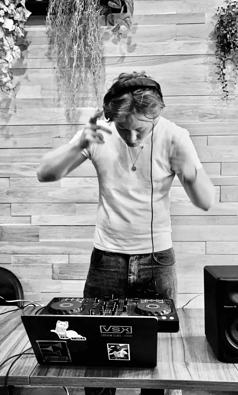 C@M in a white T-shirt, holding one headphone to his ear while mixing on a DJ controller